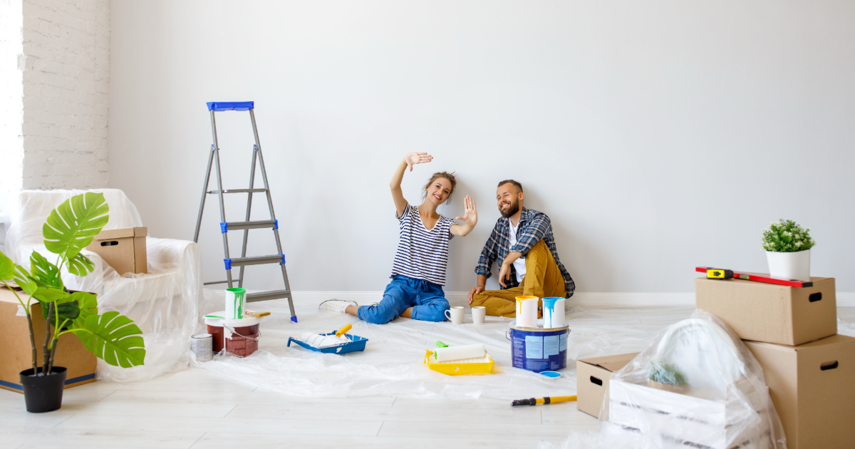 Man and woman sitting on the floor surrounded by moving boxes and ladders. 